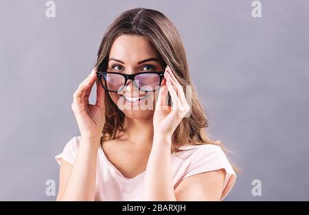 Porträt einer schönen jungen Frau, die Brille trägt und lächelt, während sie isoliert auf grauem Hintergrund steht. Fröhliches attraktives Mädchen, das eine Brille hält Stockfoto