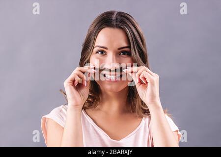 Fröhliche junge Frau in rosafarbenem T-Shirt, die einen falschen Schnurrbart ihres Haares zeigt und lächelt, während sie auf grauem Hintergrund isoliert ist. Fröhliches schönes Hüpfermädchen ho Stockfoto