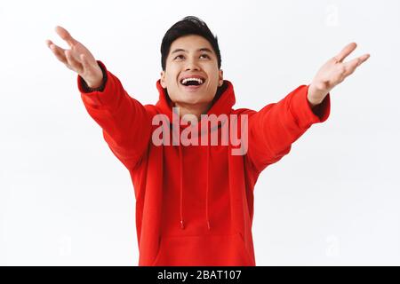 Waist-up-Porträt von verträumten glücklich jungen asiatischen Mann, trägt roten Hoodie, heben die Hände und halten Sie es bis etwas zu fangen, lächelnd zufrieden, Erfolg Stockfoto