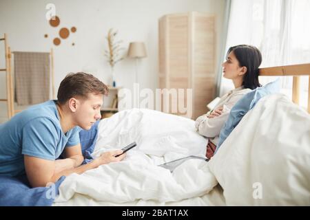 Seitenansicht des jungen Mannes mit Smartphone im Bett mit frustrierter Frau im Hintergrund, Familienkampf, Kommunikationskonzept, Stockfoto