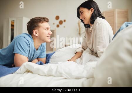 Seitenansicht Porträt des fröhlichen jungen Paares, das sich beim Sitzen auf dem Bett am Morgen betrachtet, Kopierraum Stockfoto