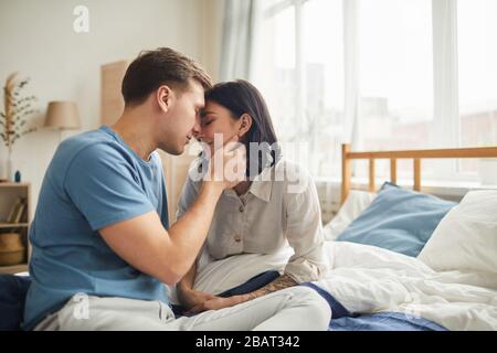 Seitenansicht Porträt des liebenden jungen Paares, das zärtlich küsst, während es morgens auf dem Bett sitzt, Kopieraum Stockfoto