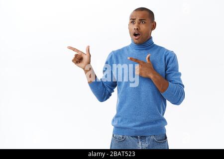 Porträt eines schockierten, sprachlosen afroamerikanischen Männchens in Turtleneck, Zeigefinger nach links, Fallbacke, besorgte und staunende, erschrocken übers Stockfoto