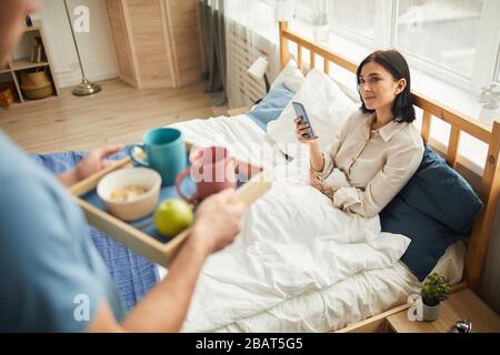 Blick in den hohen Winkel bei einem nicht erkennbaren fürsorglichen Mann, der Frühstück im Bett für junge Frau mit Smartphone, warmer Szene für Familien und Kopierraum mitbringt Stockfoto