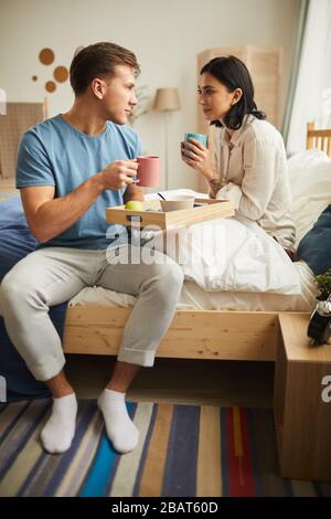 Das lange Porträt eines glücklichen jungen Paares, das Frühstück im Bett genießt und Kaffeetassen zusammenklemmt, während man sich mit Liebe ansieht Stockfoto