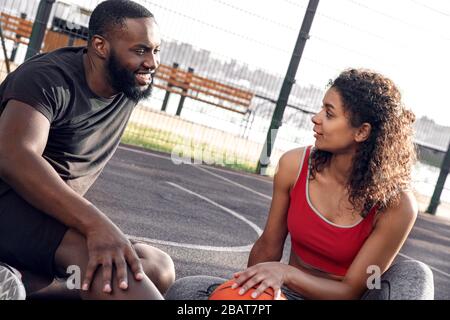 Aktivitäten Im Freien. Afrikanisches Paar sitzt mit Ball auf Basketballplatz und spricht freudige Nahaufnahme Stockfoto