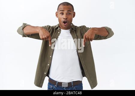 Schauen Sie hier. Porträt von amüsant, aufgeregt afroamerikanischen Mann, gute Nachrichten zu erzählen, zeigt Werbung, zeigt die Finger nach unten und schauen Kamera erstaunt Stockfoto