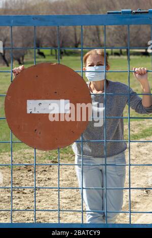 Kaukasische Frauen stehen in Schutzmaske hinter verschlossenem Tor mit rotem Stoppschild. In Quarantäne für Coronavirus Kovid-19 . Das Konzept "zu Hause bleiben" Stockfoto