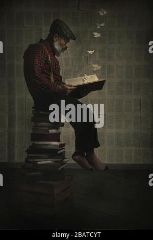 Der Mann sitzt auf einem Stapel von Büchern, die ein Buch lesen, das der Phantasie ein surreales Bild lässt. Vertikales Bild Stockfoto