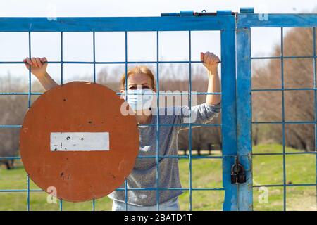 Kaukasische Frauen stehen in Schutzmaske hinter verschlossenem Tor mit rotem Stoppschild. In Quarantäne für Coronavirus Kovid-19 . Das Konzept "zu Hause bleiben" Stockfoto