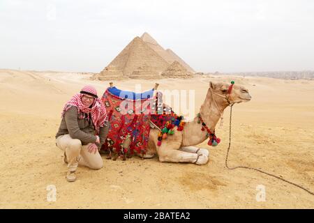 Ein Tourist kniet an einem Kamel in der Wüste und trägt eine traditionelle Nahost-Adresse beim Besuch des Giza-Plateaus in Kairo, Ägypten. Stockfoto