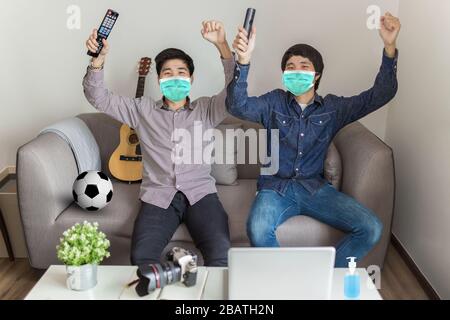 Ziel! Fröhliche Fußballfans sitzen auf dem Sofa und tragen Schutzmaske und jubeln für ein Fußballteam, das Sport-TV beobachtet. Arbeiten von zu Hause aus. Stockfoto