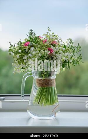 Frühling, frischer Blumenstrauß aus Liliensteinen des Tals, Rosa Rosen, blühendes Viburnum. Blumenstrauß in Glaskanne auf Fensterbank, Fensterhintergrund öffnen Stockfoto