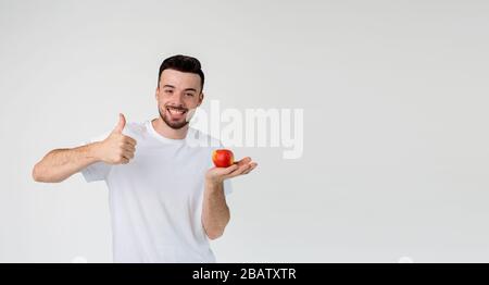 Junger Mann isoliert über Hintergrund. Fröhlicher positiver fröhlicher Kerl hält apple an der Hand. Leckeres frisches, köstliches Obst. Gesunder Lebensstil. Junge Mann zeigen große Stockfoto