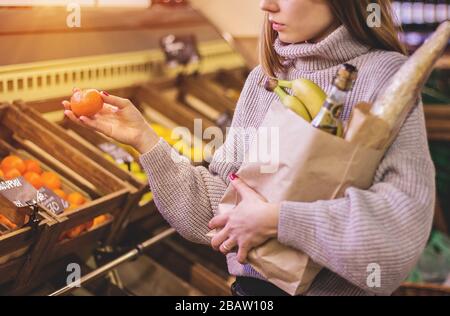 Horizontaler Schuss einer nachdenklichen Frau, die Tangerine in einer Hand hält und in einer anderen Einkaufstasche, während sie auf dem Obstmarkt steht. Schönes junges Mädchen doi Stockfoto