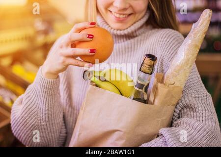 Nahaufnahme einer fröhlichen Frau, die Orange mit Essen in die Einkaufstasche legt, während sie auf dem Lebensmittelmarkt steht. Junges Mädchen steht im Supermarkt mit Beutel f. Stockfoto