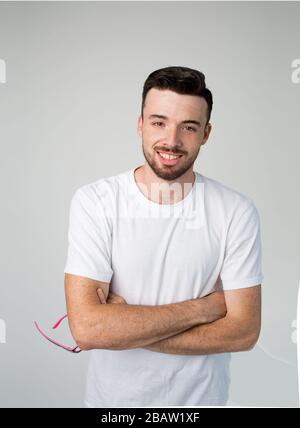 Junger Mann isoliert über Hintergrund. Porträt eines fröhlichen attraktiven, hübschen bärtigen Mannes lächelnd. Glücklicher zufriedener Kerl hält Brille in einer Hand Stockfoto