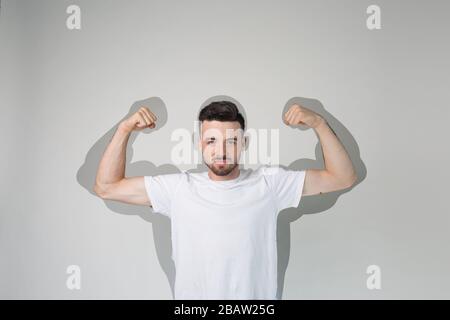 Junger Mann isoliert über Hintergrund. Starker kräftiger Kerl zeigt muskulierte Arme. Kraftvoller Schatten. Behaupten Sie, stark zu sein. Posieren allein im Studio auf der Kamera. Stockfoto