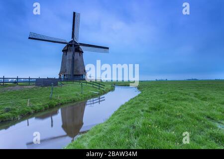 Grüne Rasenfelder und eine Windmühle spiegelten sich in der Abenddämmerung im Kanal wider. Berkmeer, Koggenland, Nordholland, Niederlande, Europa. Stockfoto