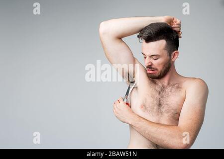 Junger Mann isoliert über Hintergrund. Nahaufnahme von Kerl, der versucht, Haare unter seine Achselhöhlen zu schneiden. Werbekonzept. Versuchen Sie, es am besten und allein zu tun. Stockfoto