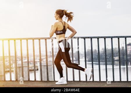 Schönes Fitness-Mädchen in stilvoller Sportswear, das über eine Brücke läuft, mit Blick auf die Stadt im Hintergrund. Schlanke junge Sportlerin beim Joggen in der Stadt. Attraktiv Stockfoto
