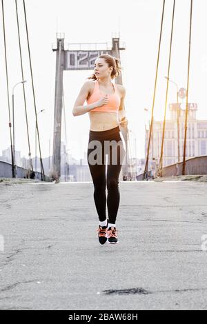 Attraktive junge Sportlerin im stilvollen Sport trägt Joggen über Brücke mit urbaner Sicht auf den Hintergrund. Wunderschönes Fitness-Mädchen, das auf der Straße in der Stadt läuft. Stockfoto