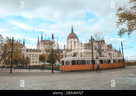 Budapest, Ungarn - 6. November 2019: Öffentliche gelbe Straßenbahn vor dem ungarischen Parlamentsgebäude. Menschen auf der Straße. Hauptstadt des öffentlichen Personennahverkehrs. Stadtverkehr. Touristenattraktion. Stockfoto