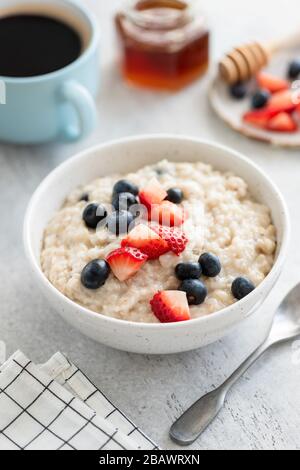 Haferbrei mit Erdbeeren und Blaubeeren auf grauem Betontisch serviert mit einer Tasse schwarzen amerikanischen Kaffee und rohem Honig im Mixbecher. Gesundes BRE Stockfoto