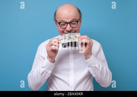 Ein älterer Kaukasusmann in Gläsern hält eine Dollar-Banknote und Schnüffeln Stockfoto