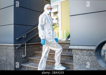 Junge Erwachsene Frau mit Gesichtsmaske und biologischem Hazmat-Schutzanzug und Handschuhen, die in der Nähe des Gebäudeeingangs im Freien laufen. Mutter geht einkaufen Stockfoto