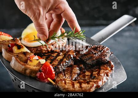 Grillplatte. Verschiedene leckere gegrilltes Fleisch mit Gemüse. Gegrilltes gemischtes Fleisch mit Pfeffersauce und Gemüse. Die Hände des Küchenchefs verteilen Fleisch Stockfoto