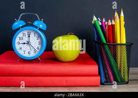 Der blaue Wecker sowie mehrfarbige Bleistifte stehen auf einem Stapel roter Bücher, neben einem apfel Stockfoto