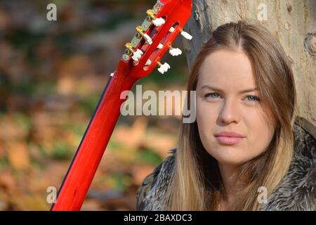 Portrait des jungen, schönen, blauäugigen Mädchens mit roter Akustikgitarre unter großem planetreem, natürlichem Look Stockfoto