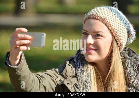 Süßes, junges Mädchen mit Wollmütze, das selfie an einem sonnigen Herbsttag per Handy im Freien nimmt Stockfoto