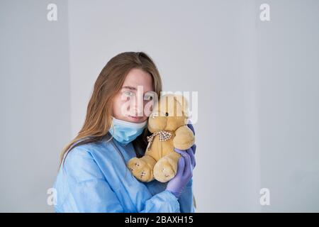 Ein Nahporträt eines Arztes, der ein Operationssheater-Outfit trägt und einen Teddybären lötet - isoliert auf blauem Grund. Konzept der Zärtlichkeit an Stockfoto