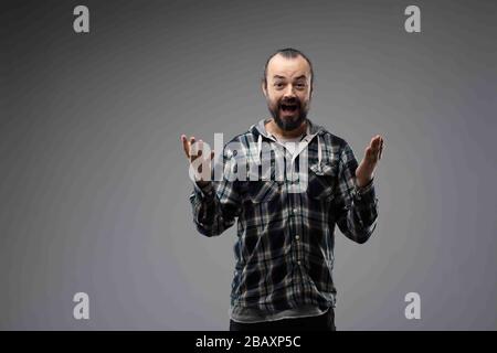 Glücklicher bärtiger Mann im karierten Hemd, der überraschte Geste zeigt, mit den Händen vor ihm, mit Blick auf die Kamera. Halblanges Vorderporträt gegen Grau Stockfoto