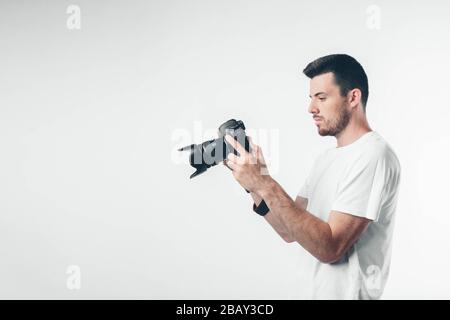 Junger brünette Mann mit Bart in weißem T-Shirt. Guy hält professionelle Kamera mit großem Objektiv in der Hand. Er ist ernst und konzentriert Stockfoto