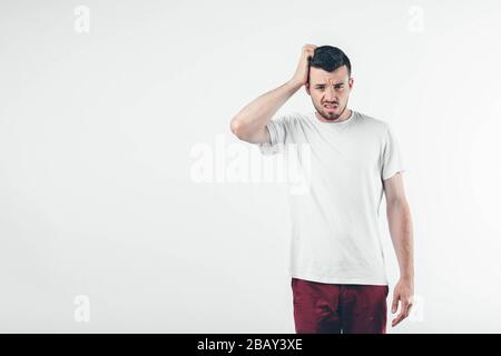 Junger brünette Mann mit Bart in weißem T-Shirt. Kerl onfusioniert und verärgert. Mit der rechten Hand kratzt er sich den Kopf. Isoliert über Licht Stockfoto