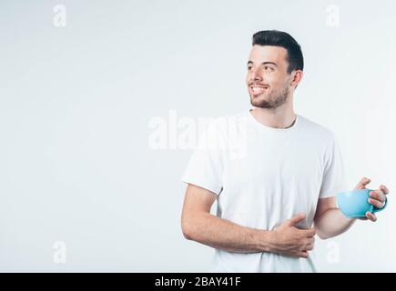 Junger brünette Mann mit Bart in weißem T-Shirt. Guy lächelt eine Hand auf seinem Bauch, die zweite hält den blauen Becher. Isoliert über hellem Hintergrund. Stockfoto