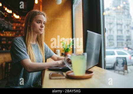 Die junge Blonde in einem Café arbeitet mit einem Laptop. Auf dem Tisch steht eine Tasse grünen Tee. Stockfoto