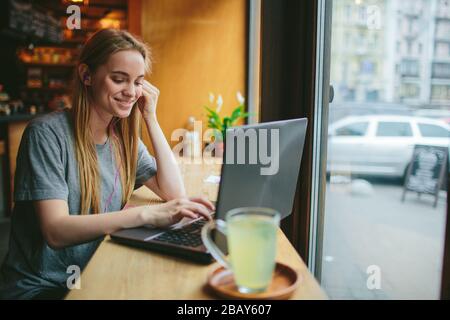 Die junge Blonde in einem Café arbeitet mit einem Laptop und hört Musik. Auf dem Tisch steht eine Tasse grünen Tee. Stockfoto