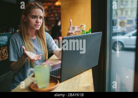 Die junge Blonde in einem Café arbeitet mit einem Laptop. Sie ist verwirrt. Auf dem Tisch steht eine Tasse grünen Tee. Stockfoto