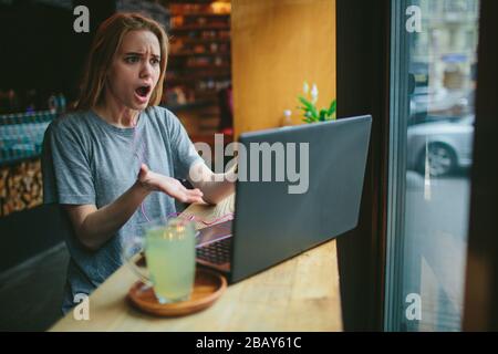 Die junge Blonde in einem Café arbeitet mit einem Laptop. Sie ist verwirrt und überrascht. Auf dem Tisch steht eine Tasse grünen Tee. Stockfoto