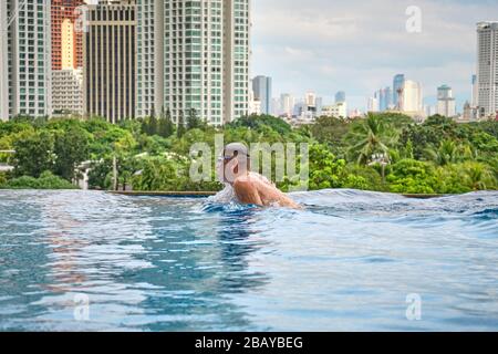 Ein Mann schwimmt einen Schmetterling in einem Pool auf dem Dach eines Luxushotels. Blick auf die Stadt Manila vom Pool des luxuriösen Fünf-Sterne-Discovery Primea Hot Stockfoto