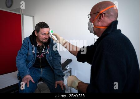München, Deutschland. März 2020. Arzt Hans-Gerd Schmiz misst die Temperatur mit einem kontaktlosen klinischen Thermometer während seiner Sprechstunden im Fliednerhaus der Diakonie bei Thorsten Böhme. Obdachlose sind besonders von der Corona-Pandemie betroffen. Das Hilfesystem zerfällt zunehmend, gleichzeitig müssen die oft immungeschwächten Menschen vor allem eine Infektion fürchten. Credit: Bernd Thissen / dpa / Alamy Live News Stockfoto
