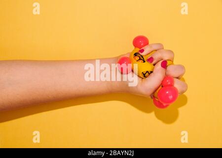 Weibliche Hand mit rotem Nagellack, die einen bunten roten und gelben Spielzeugball eines Kindes über einen orangefarbenen Hintergrund mit Schatten- und Kopierraum wirft Stockfoto