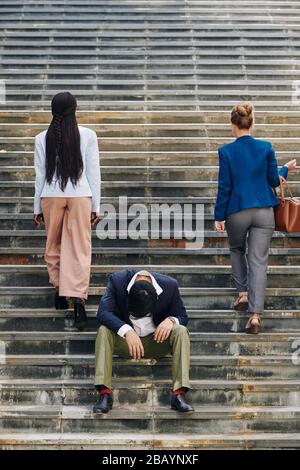 Betont frustrierter arbeitsloser junger Mann, der auf Stufen sitzt, wenn junge Geschäftsfrauen die Treppe hinauf gehen Stockfoto