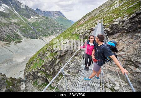 Von oben Blick auf liebendes Paar mit Rucksäcken auf Hängebrücke, schöne Berglandschaft im Hintergrund. Wilde Natur mit fantastischer Aussicht. Sport Tourismus in den Alpen. Stockfoto