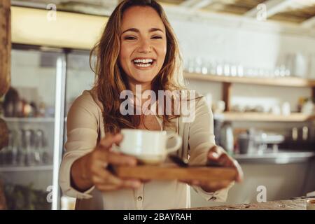Lächelnde Frau, die hinter dem Ladentisch arbeitet. Fröhliche junge Kellnerin, die dem Kunden Kaffee im Café serviert. Stockfoto
