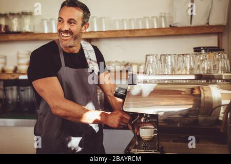 Barista, männlich, bereitet Espresso im Café vor, der sieht weg und lächelt. Mann in der Schürze, der Kaffee mit einer Kaffeemaschine im Café zubereiten kann. Stockfoto
